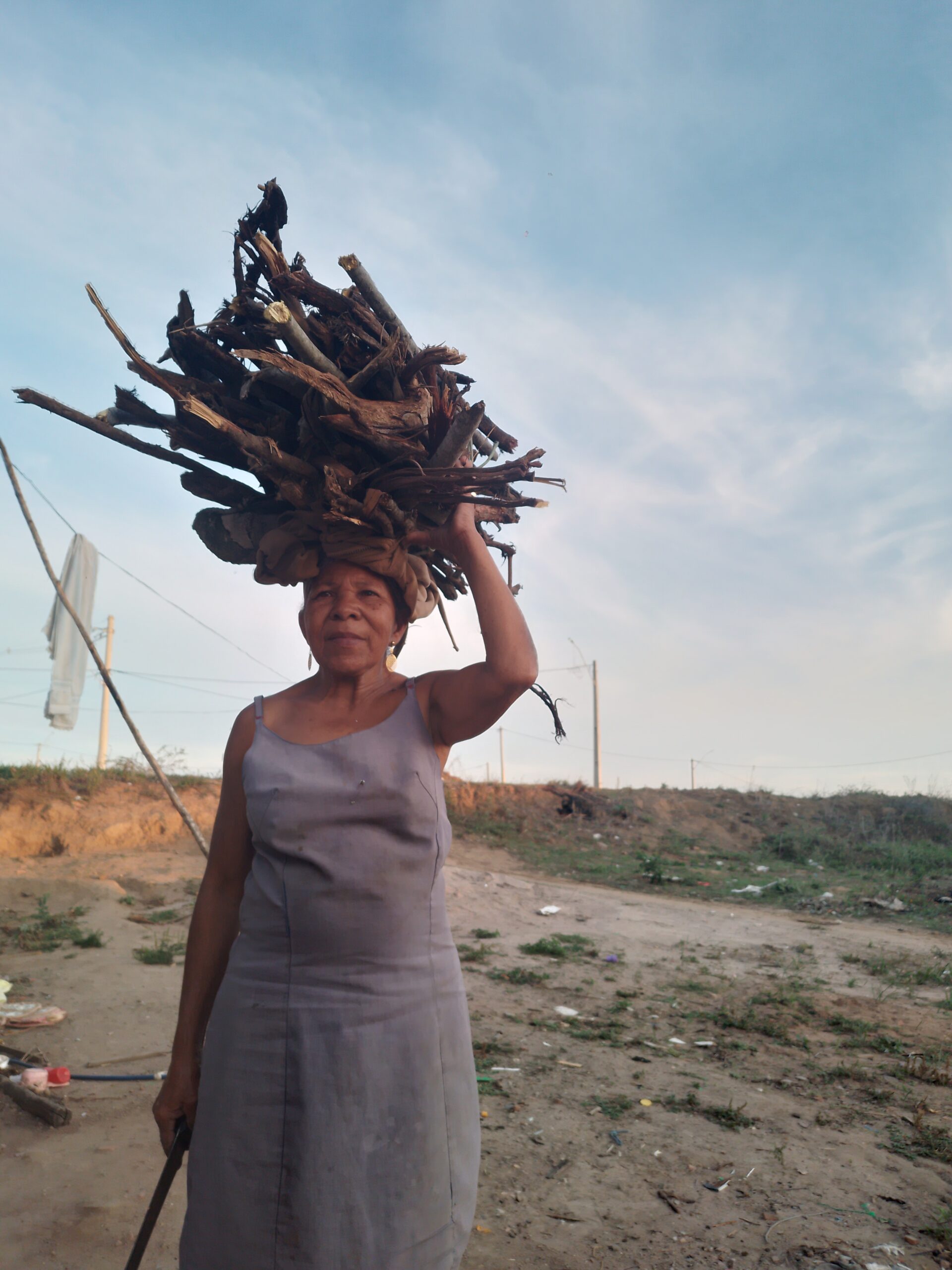 Uma senhora negra de pele clara e cabelos grisalhos aparece em pé em um ambiente rural. Ela está usando um vestido cinza e equilibrando na cabeça um grande feixe de galhos secos. Ela segura uma ferramenta com uma das mãos, e o cenário ao fundo é de um terreno com vegetação rala e um céu claro ao entardecer.